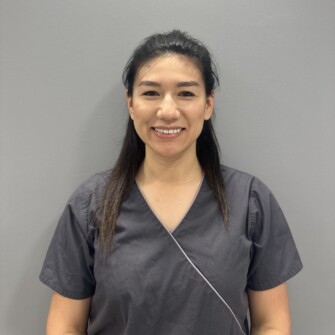 A female dental professional wearing a grey uniform and smiling at the camera against a plain background.