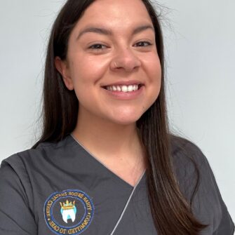 A dental professional smiling while wearing grey scrubs with an emblem on the chest.