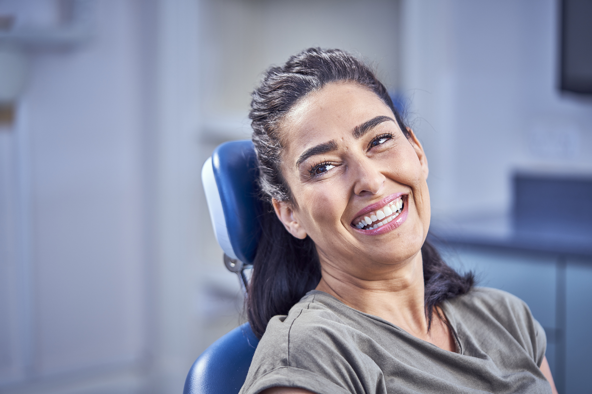 A woman with long dark hair smiling while seated in a dental chair at a dental practice.