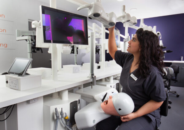 Instructor demonstrating a dental training simulation with a mannequin and monitor in a clinical setting.