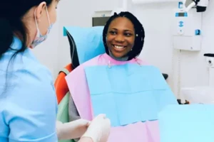 A patient sitting in a dental chair wearing a bib while smiling at a dental assistant in a clinic. A patient sitting in a dental chair wearing a bib while smiling at a dental assistant in a clinic.