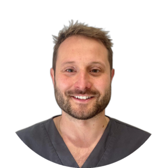 Portrait of a dental professional in grey scrubs with brown hair and a trimmed beard, smiling against a clean white studio background