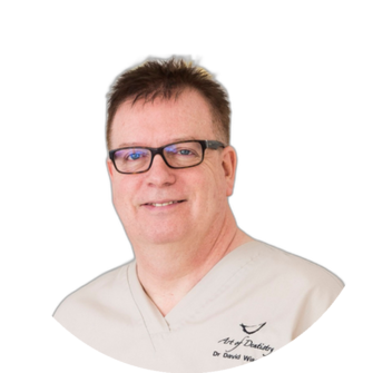 David Wiseman wearing beige scrubs and dark-framed glasses, smiling softly against a white background.