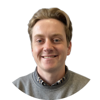 Henry Cairns portrait of a young man with blond hair, wearing a grey sweater over a check shirt, smiling against a white background