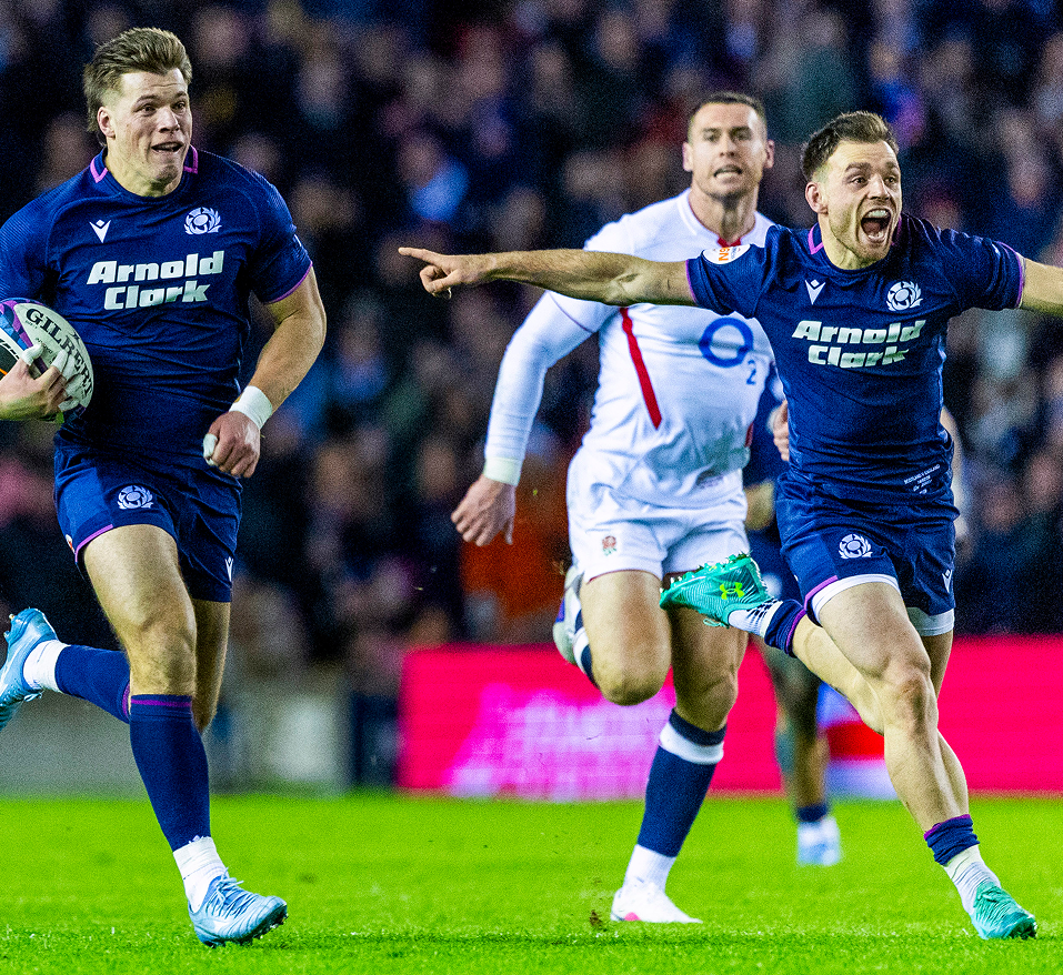 Arnold Clark rugby players in blue kits sprint across a green pitch, one player holding a ball as teammates vie for space