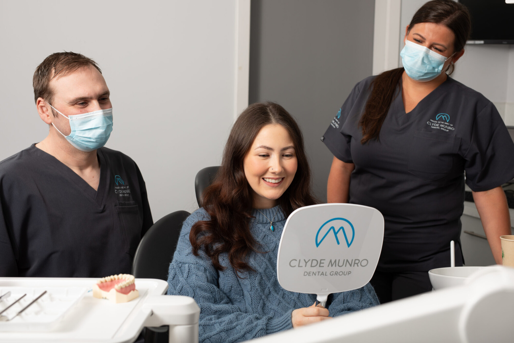 A smiling teenage patient in a blue jumper sits with masked Clyde Munro dental staff during an orthodontic consultation.