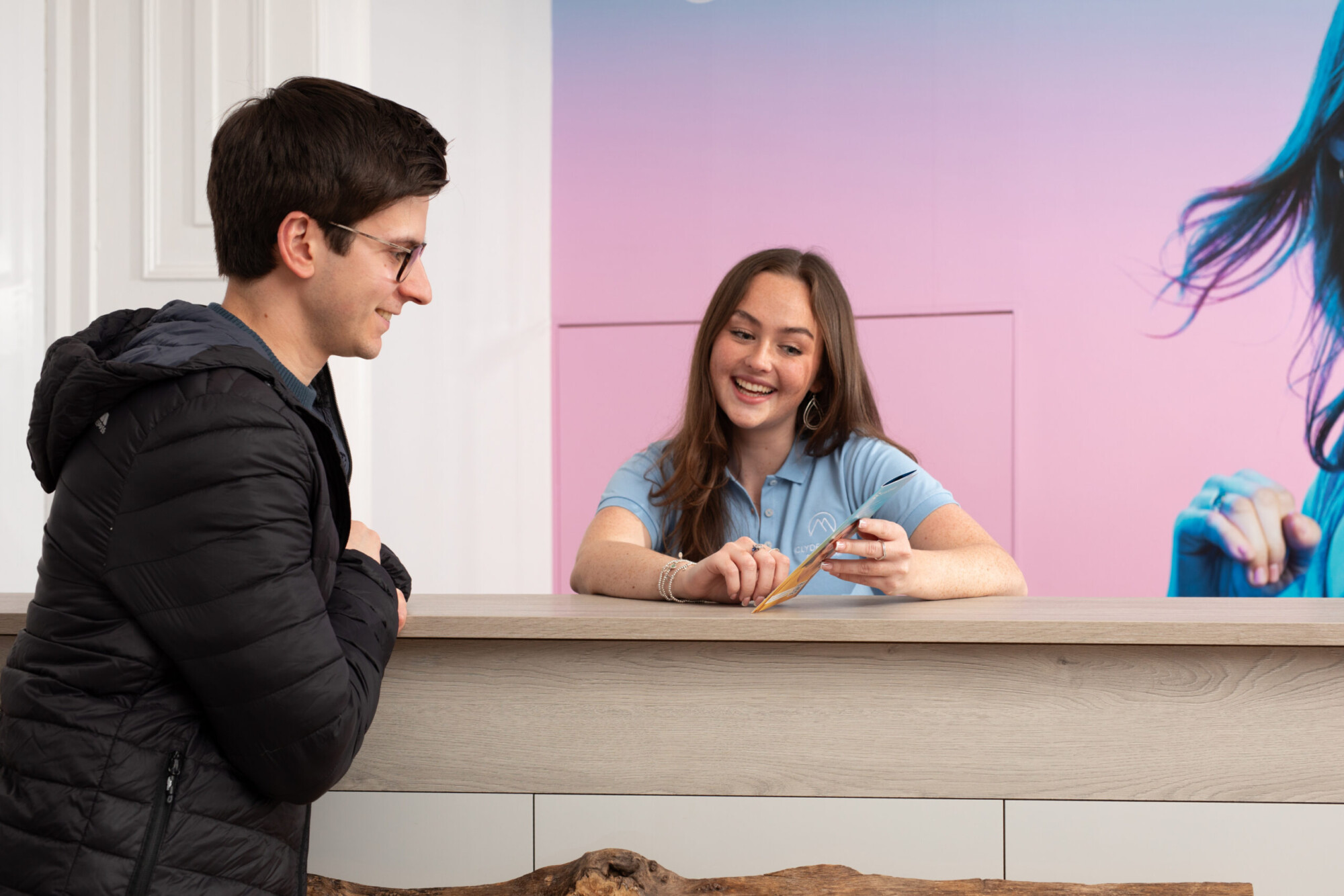 A smiling Clyde Munro team member in a blue polo speaks with a young man at the reception desk beside a pink wall.