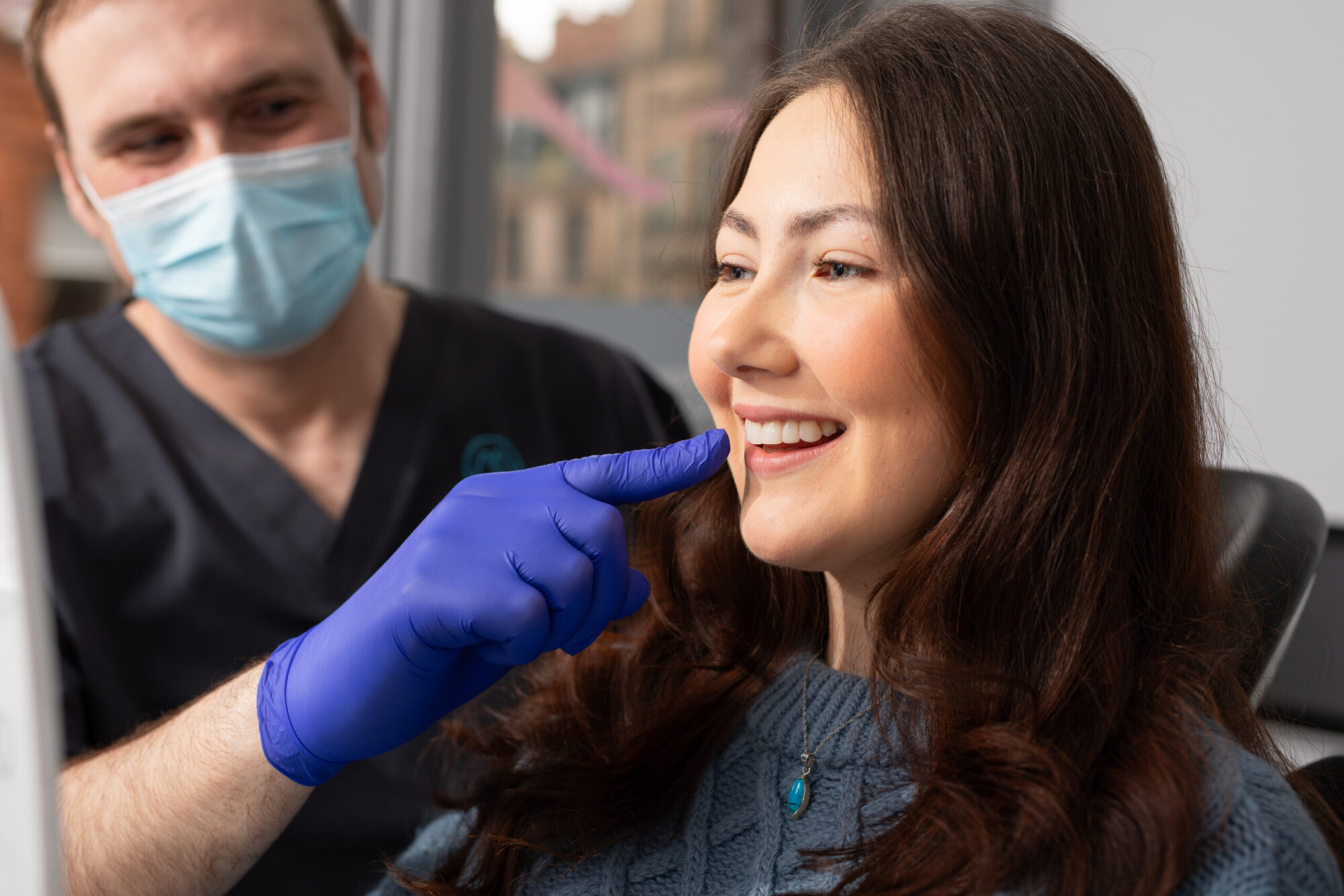 A masked clinician in blue gloves points to a smiling woman’s teeth during a dental consultation.