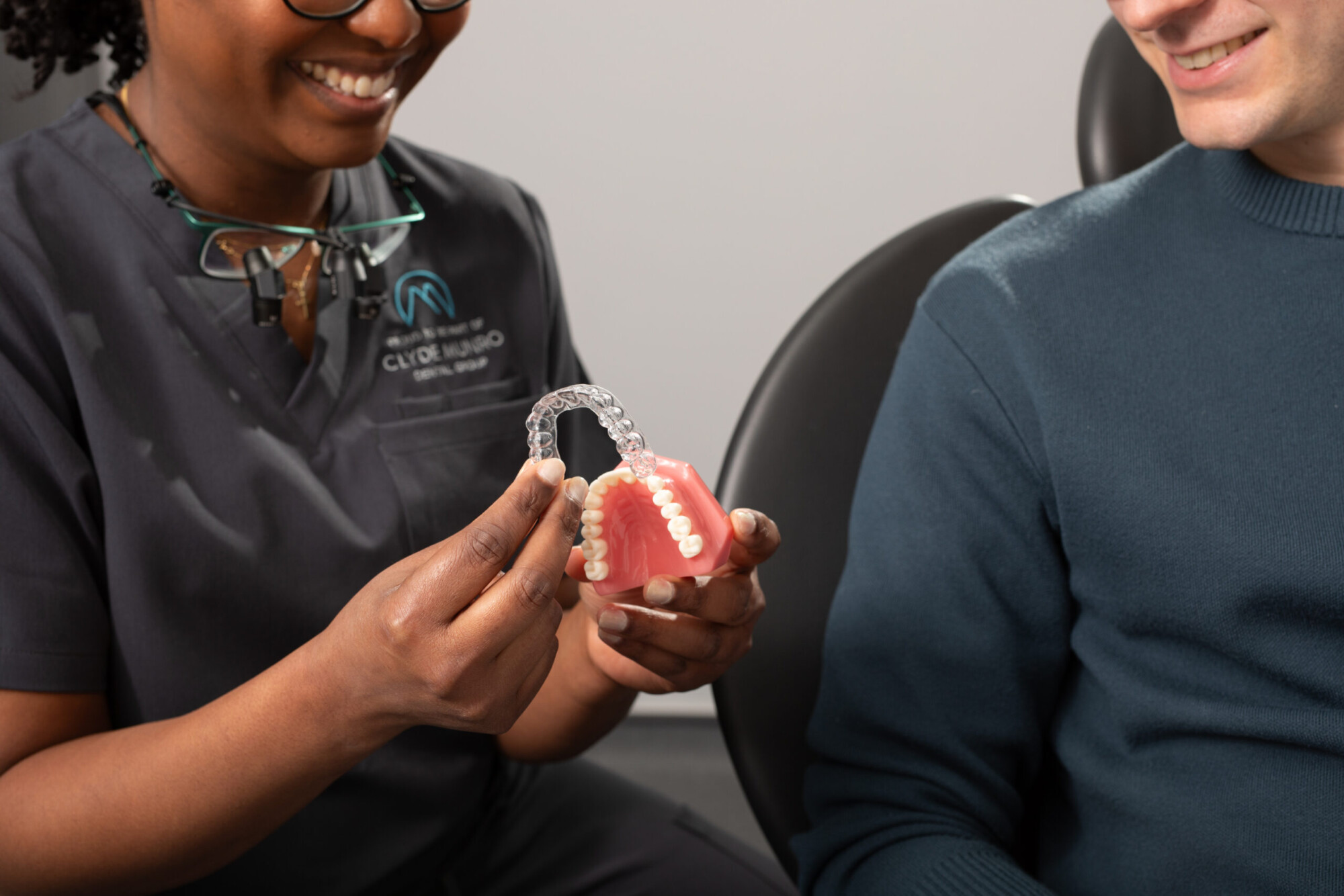 A Clyde Munro clinician shows a clear aligner over a dental model while talking with a smiling patient.