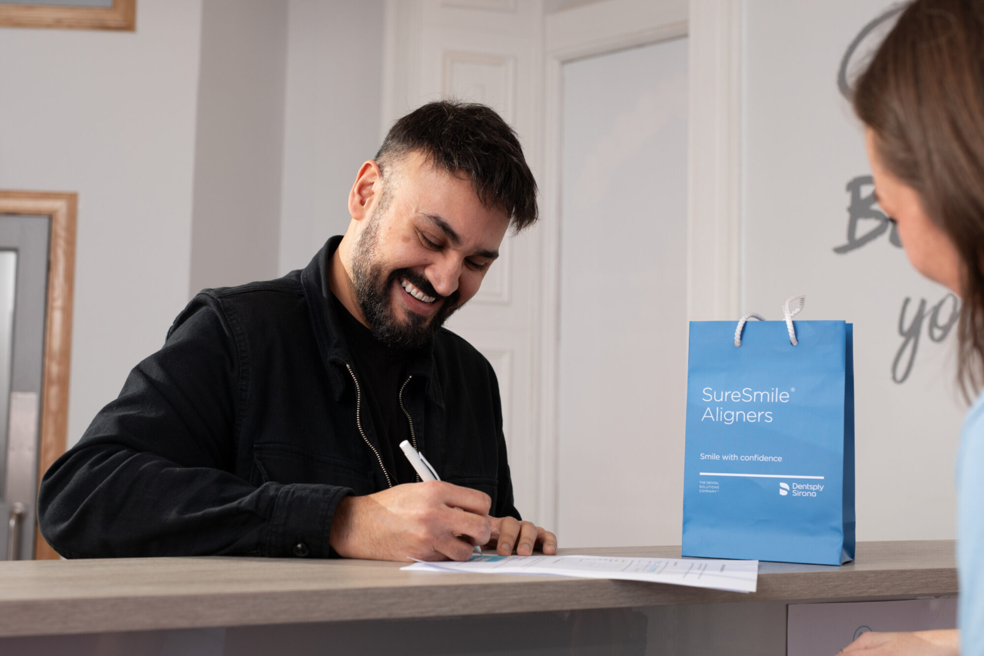 A smiling man signs paperwork beside a blue SureSmile Aligners bag during a consultation at Clyde Munro dental practice.