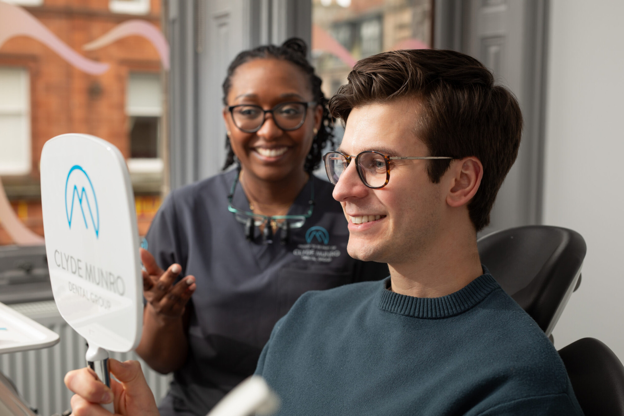 A smiling patient holds a Clyde Munro dental mirror while a clinician stands behind him in a modern treatment room.
