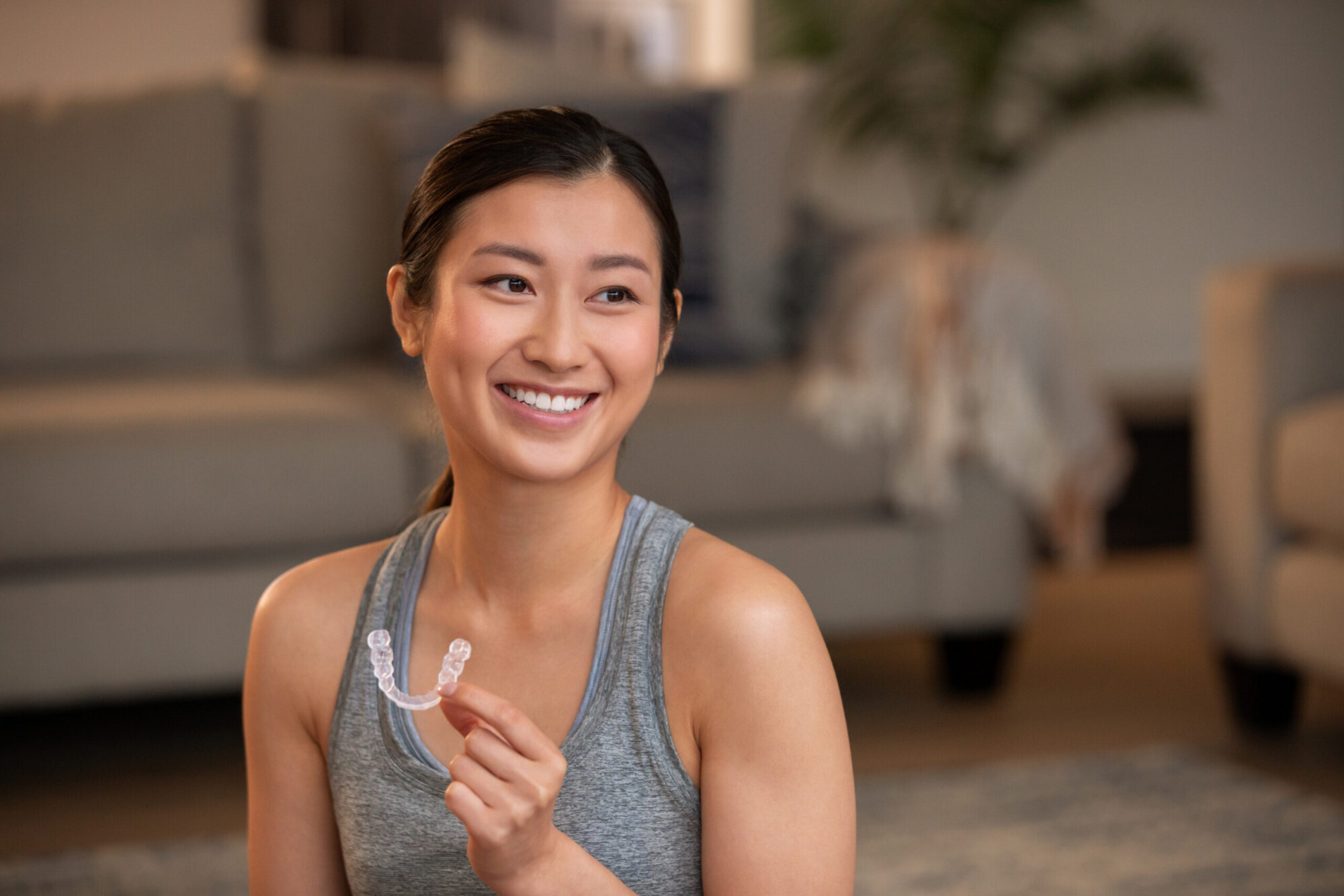 A smiling woman in a grey vest holds a clear Invisalign aligner between her fingers in a softly lit home setting.