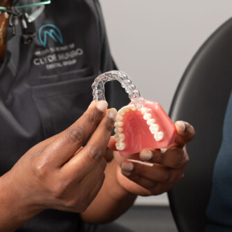 A Clyde Munro Dental Group clinician holds a clear aligner above a pink dental model while discussing tooth alignment with a patient nearby.