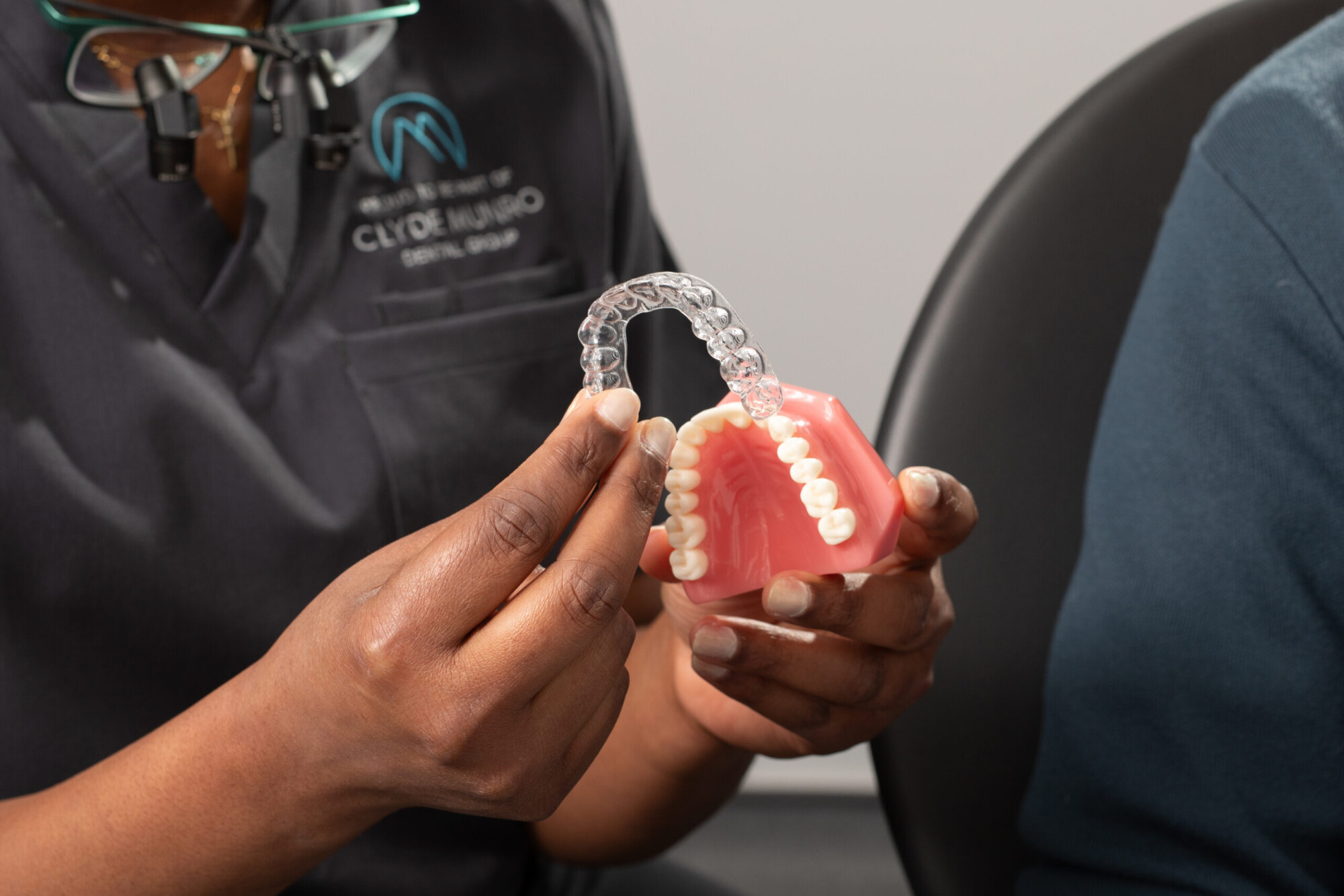 A dentist in Clyde Munro uniform holds a clear aligner above a dental model while examining the fit during a consultation.