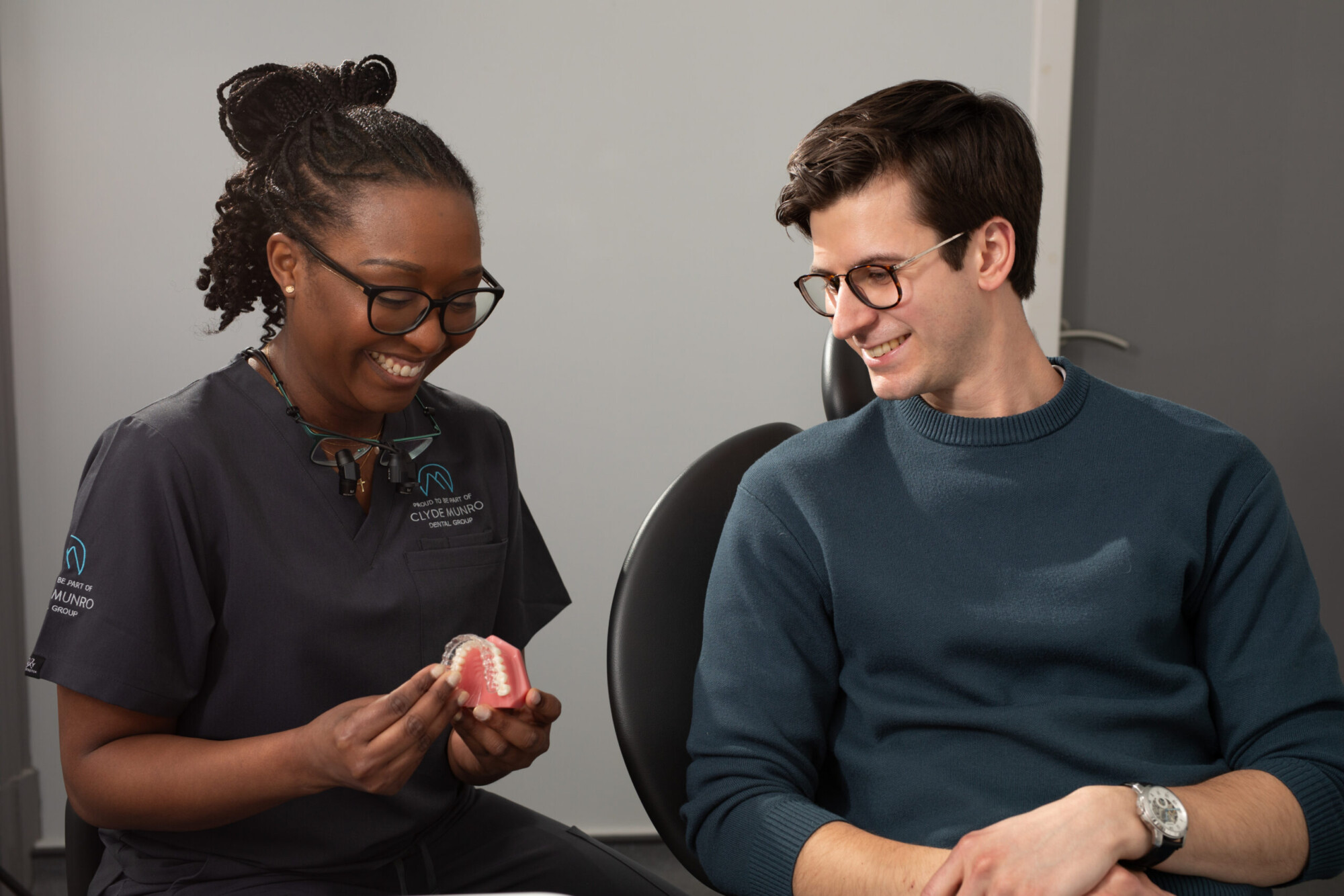 A Clyde Munro clinician in black scrubs shows a clear aligner model to a smiling patient wearing glasses during an orthodontic consultation.
