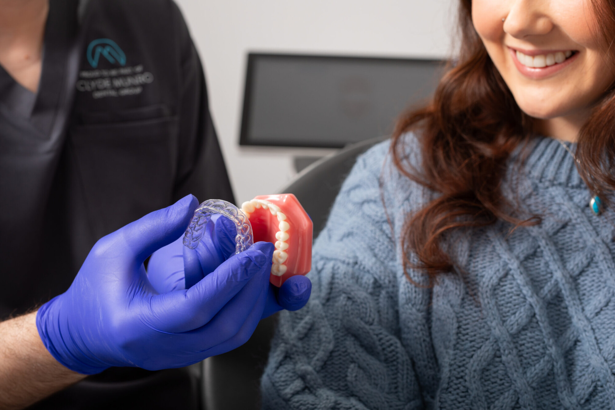A clinician in blue gloves shows a clear aligner beside a dental model while a smiling patient looks on at Clyde Munro.