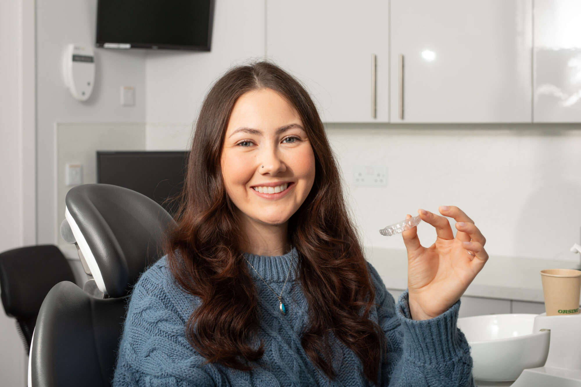 A smiling young woman in a dental clinic holds a clear aligner while seated beside the treatment chair.