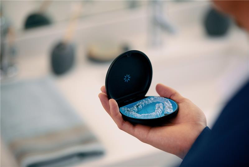 A hand holds an open Invisalign case with a clear aligner tray inside in a bright dental setting.