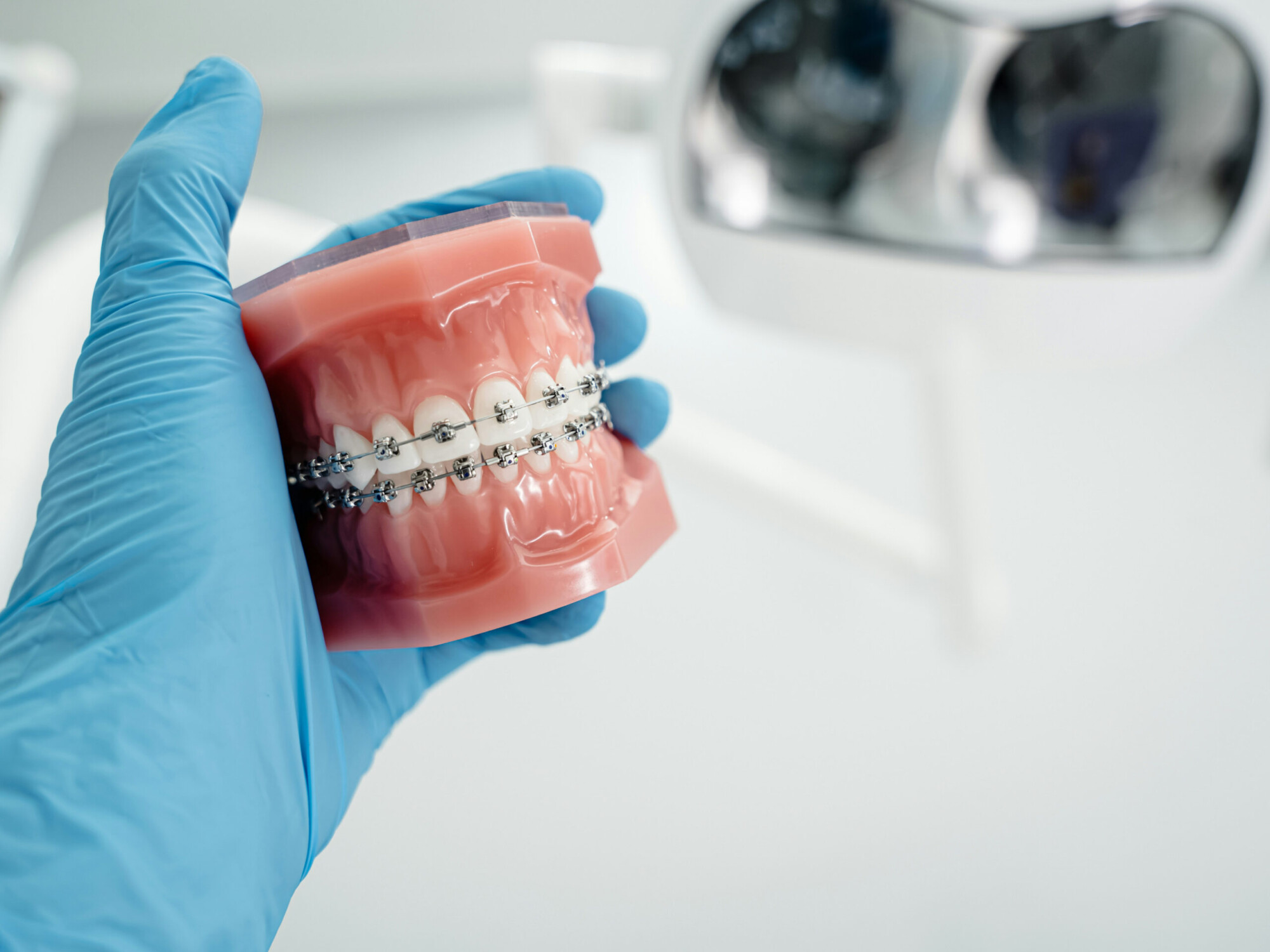 A blue-gloved hand holds a dental model fitted with metal fixed braces, with a blurred dental mirror in the background.