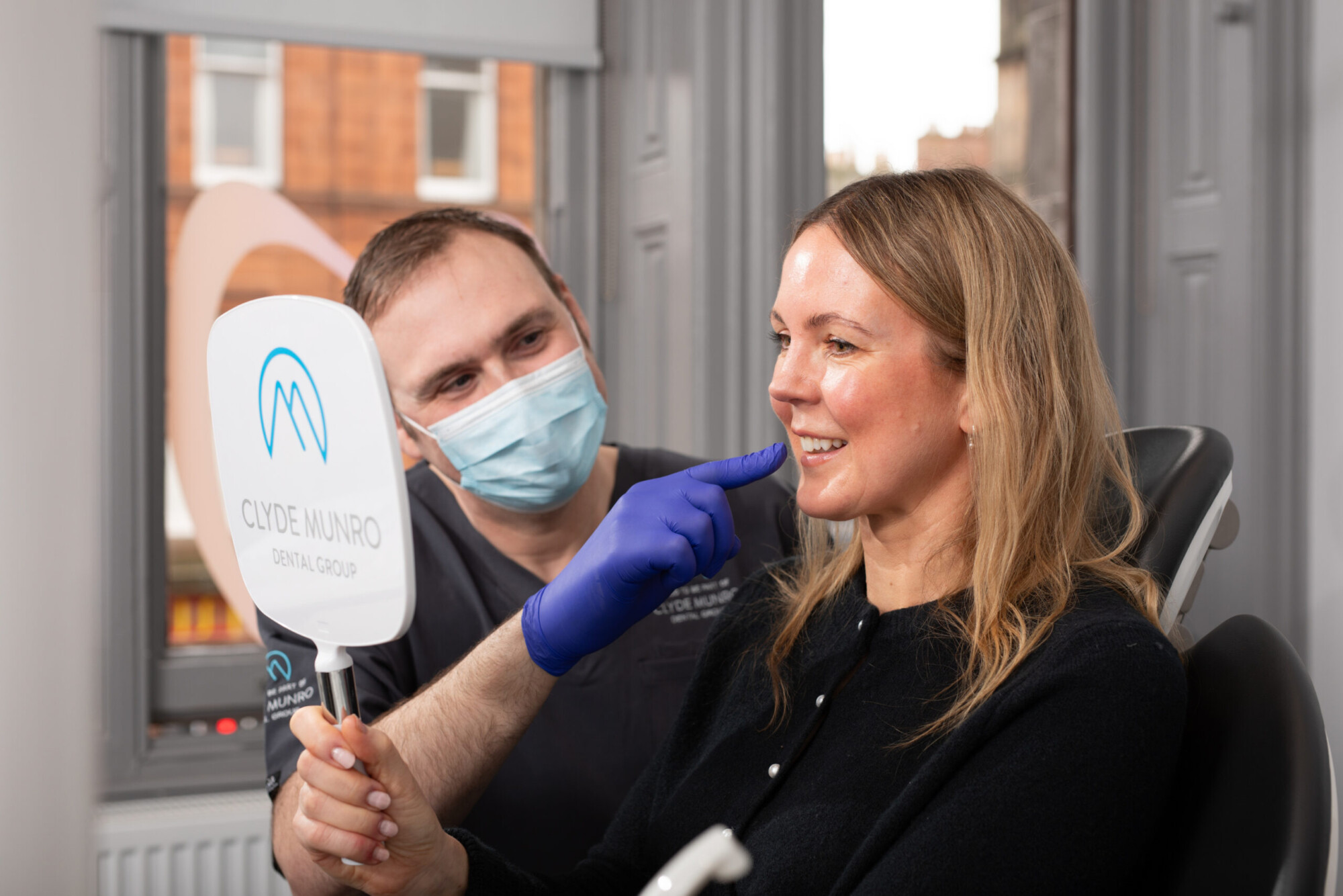 A dentist in blue gloves and a face mask shows a patient her smile with a handheld mirror at Clyde Munro Dental Group.